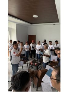 Foto 4. Latihan koor perdana di ruang utama Lakeside Haus Balige, Jumat 24 Juli 2026 pukul 10.30 WIB, tak lama setelah rombongan tiba dari Bandara Silangit. Dirigen Amang Parlis Sinaga memimpin dari depan sementara para peserta membaca partitur lagu yang akan dibawakan pada Ibadah Minggu di HKBP Balige.
