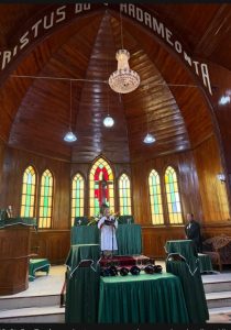 Foto 19. St. Eva Tambunan bertugas maragenda — membacakan liturgi ibadah — pada Ibadah Minggu di HKBP Balige, 26 Juli 2026. Di dinding melengkung tampak tulisan "Kristus do Pardameonta".