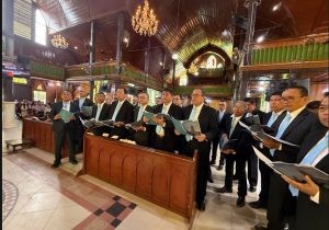 Foto 21. Punguan Koor Ama HKBP Slipi membawakan lagu pujian pada Ibadah Minggu di HKBP Balige, 26 Juli 2026. Papan pengumuman di sisi kiri ruangan menampilkan nama kelompok koor yang sedang tampil.
