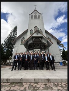 Foto 22. Rombongan Punguan Koor Ama HKBP Slipi berfoto bersama di halaman depan Gereja HKBP Balige, seusai Ibadah Minggu, 26 Juli 2026. Bangunan gereja kayu bergaya gotik dengan menara menjulang dan jendela kaca patri ini merupakan salah satu penanda sejarah kekristenan di Tanah Batak. Tampak pula pelayan gereja setempat berdiri bersama rombongan.